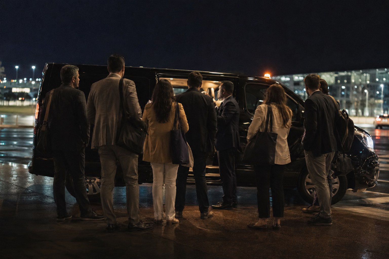 Grupo de executivos brasileiros de costas, com bolsas e pastas, embarcando numa van preta à noite enquanto um deles fala com o motorista na porta lateral